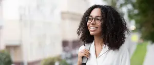 A woman with curly hair is smiling and looking to the right. She is wearing glasses and a white shirt. Behind her is a blurry background of a building with a parked car and a tree.