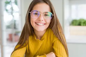 A girl with brown hair and glasses smiling and sitting in a room with glass windows.