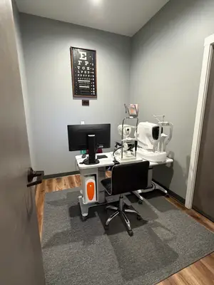 The interior of an examination room in a medical clinic with an examination table, a computer monitor, a chair, and a carpeted floor, and a wall-mounted eye chart. There is also a door with a handle and a power outlet on the wall.