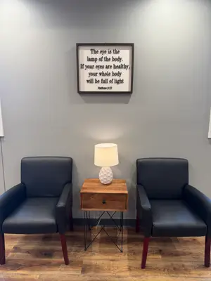 Two black chairs are placed on a wooden floor in a room with a gray wall, a wooden table with a lamp, and a picture frame with a motivational quote. The picture frame is mounted on the wall, and the quote reads, 'The eye is the lamp of the body. If your eyes are healthy, your whole body will be full of light. Matthew 6:22'. The room is well-lit, and the wooden floor reflects the light.
