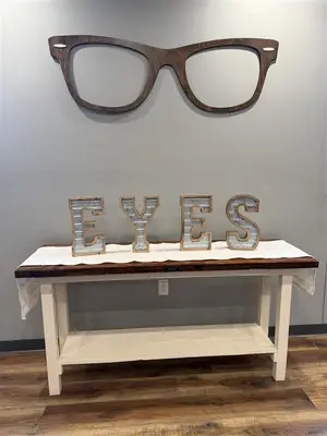 A wooden table with a white cloth on it and a sign that reads EYES in front of a gray wall with a wooden frame of eyeglasses