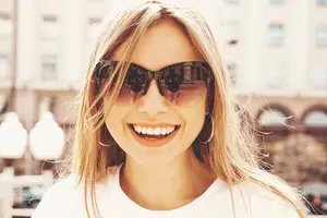 A woman with blonde hair is smiling and posing for a photo. She is wearing sunglasses and a white shirt. Behind her is a blurry view of a street with streetlights and buildings.