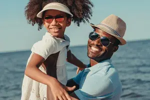 An adult male and a young girl are standing on the beach and smiling at the camera