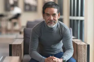 A man sitting on a couch inside a room with wooden accents, wearing a gray long-sleeve shirt and blue pants.