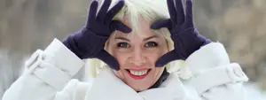 A woman with blonde hair is wearing a white coat and purple gloves, smiling and posing for a photo in a snowy area.