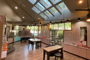 An interior view of an office with a wooden floor, a ceiling with skylights, glass windows, a desk, chairs, a cabinet, and lamps hanging from the ceiling. A person is standing near the desk with a monitor, and there is a staircase with flowers on the left side. There is also a mirror with a red sign on the right side.
