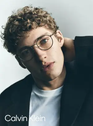A close-up of a man with curly hair wearing a white shirt, a black blazer, and glasses with a Calvin Klein logo. He is posing for a photo in front of a white background.