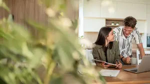 A couple sitting in a kitchen area, the woman is looking at a paper while the man is smiling