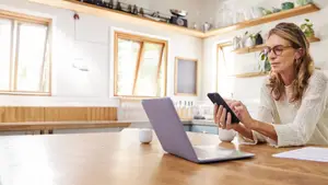 A woman sits at a desk in her kitchen, holding a smartphone and a laptop, with a cup and a document nearby, suggesting she is working or browsing online.