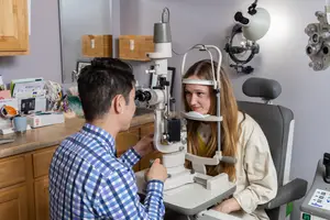 A woman with long blonde hair sits in a chair and has her eyes examined by a man in a blue and white checkered shirt