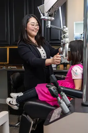 A woman with glasses is adjusting a medical machine while sitting a girl in a pink dress next to her.