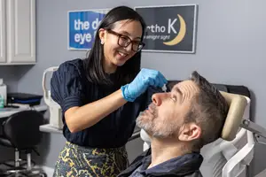 Woman wearing blue gloves administers eye drops to a man in an office setting