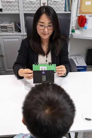 A woman wearing glasses is showing a green and black folder with a purple sticker to a boy sitting in front of her.