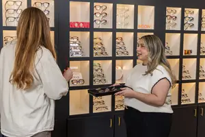 Two women are standing in front of a display case with glasses, one holding a black tray of glasses while the other looks at them, with a wall of glass cases in the background displaying different glasses and the word 'WOOW' on them.