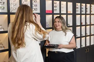 A smiling woman in a white blouse tries on glasses in an optical shop, while another woman holds a tray of eyeglasses.