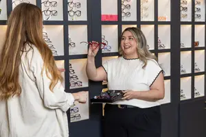 Two women smiling at each other, one holding a red-framed pair of glasses, inside a shop with shelves of glasses in the background.