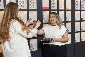 Two women are standing inside a store with many glass cases displaying different pairs of eyeglasses. One woman is holding a pair of eyeglasses while the other woman is holding a box with a pair of eyeglasses inside.