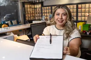 Woman holding a clipboard with a pen, smiling and looking at the camera in a room with white walls and a white counter, surrounded by furniture and a large glass window