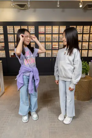 Two girls standing in front of a display case of glasses, one wearing a shirt with a cartoon character and the other in a gray sweatshirt, both smiling and holding glasses