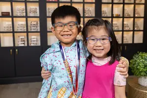 A young boy and girl wearing glasses are smiling and posing for a photo in a room with a display cabinet behind them.