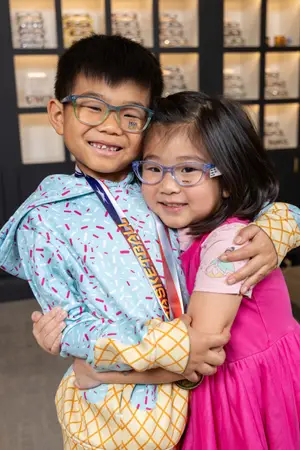 A boy wearing a medal and a girl wearing glasses are hugging each other in front of a glass cabinet