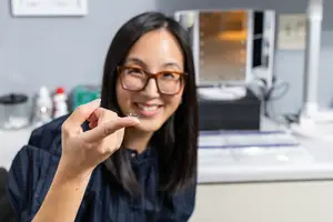 An adult woman holds a contact lens in her hand while smiling in a lab setting