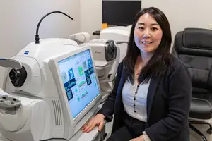 A woman sitting next to an ophthalmoscope machine in an office room