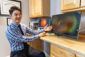 A smiling man in a checkered shirt and tie shows a monitor with a retinal image to someone in a home office setting.