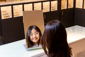 A girl with glasses is looking at her reflection in a mirror in a room with cabinets and shelves with glasses frames