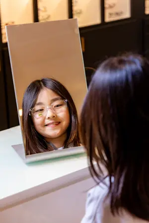 A girl with glasses is looking at her reflection in a mirror while smiling in an indoor area.