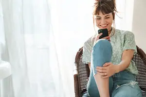 A woman is sitting on a chair and holding a cellphone in her hand while smiling. She is wearing a shirt and jeans. She is probably listening to music. Behind her is a white curtain. On the left side, there is a white object that seems to be a sink.