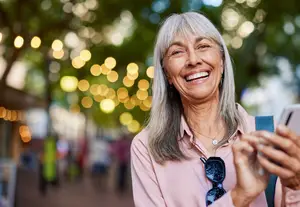 An older woman with white hair is standing on the street, smiling, and looking at her phone.