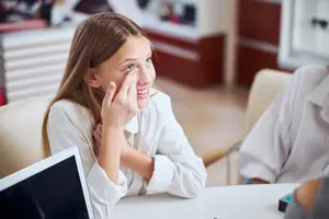A young girl is sitting in a chair, smiling and looking at her laptop, with her hand on her face.