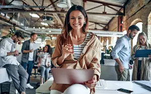 A woman is sitting on a chair in an office and holding a laptop, a pen, and a document while smiling and looking at the camera. Behind her, there are people standing and sitting on chairs. One of them is holding a cell phone. There are plants and lamps hanging from the ceiling. The wall is made of bricks and has a window with a view of the outside.