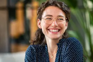 A smiling woman with glasses and a polka dot shirt standing in a room with a blurry background.