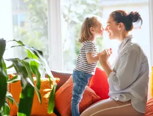 A woman and a young girl are standing in front of a window, smiling and looking at each other. The woman is holding the girl's hand. They are sitting on a couch with orange and red pillows. The woman has a ponytail, and the girl has a ponytail. A potted plant is on the left side, and a building is outside the window.