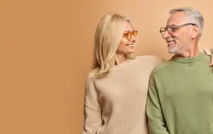 An elderly man and woman standing together, smiling and looking at each other, against a light brown background