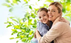 A woman and a girl are smiling and hugging each other in front of a tree with green leaves.