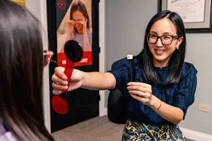 A woman is sitting in a chair holding a red object in her hand and is smiling at the camera. Another woman is standing in front of her and is also holding a red object. Behind them is a poster of a woman with glasses and a door.