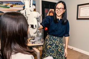 A smiling woman wearing glasses is checking the eye of a patient in an optometrist office