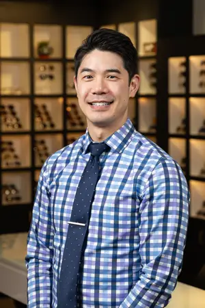 A smiling man in a blue checkered shirt stands in front of a wall with glass shelves displaying glasses