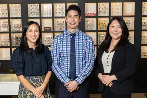 Three smiling people stand in front of a wall with glasses displayed on shelves