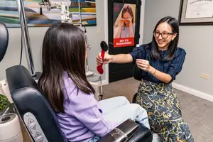 A woman and a young girl are in an optical clinic, with the woman demonstrating something to the girl while the girl looks at her with a curious expression. The room has a black door with a poster on it, and a white wall with a certificate and a painting.