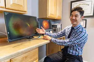 A smiling man in a blue and white checkered shirt is sitting at a desk in front of a computer screen showing an eye image, with cabinets and framed certificates in the background.