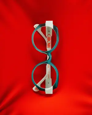 A pair of Etnia Barcelona﻿ Chroma eyeglasses with teal frames and white lenses against a red background