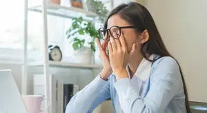 A woman in a blue sweater sitting at a desk, covering her eyes with her hands and looking away.
