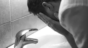 A man with his face in his hands standing at a sink with water running