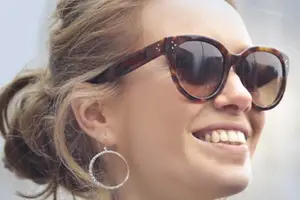A smiling woman with brown hair and sunglasses is wearing a silver hoop earring and is looking at something outside.