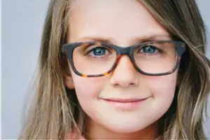 A young girl with brown hair is wearing glasses and smiling at the camera in a photo studio.