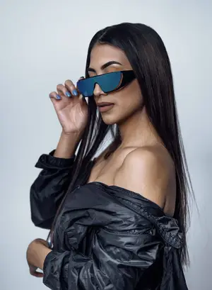 A woman wearing sunglasses with a black jacket and blue nails is posing for a photo with a white background
